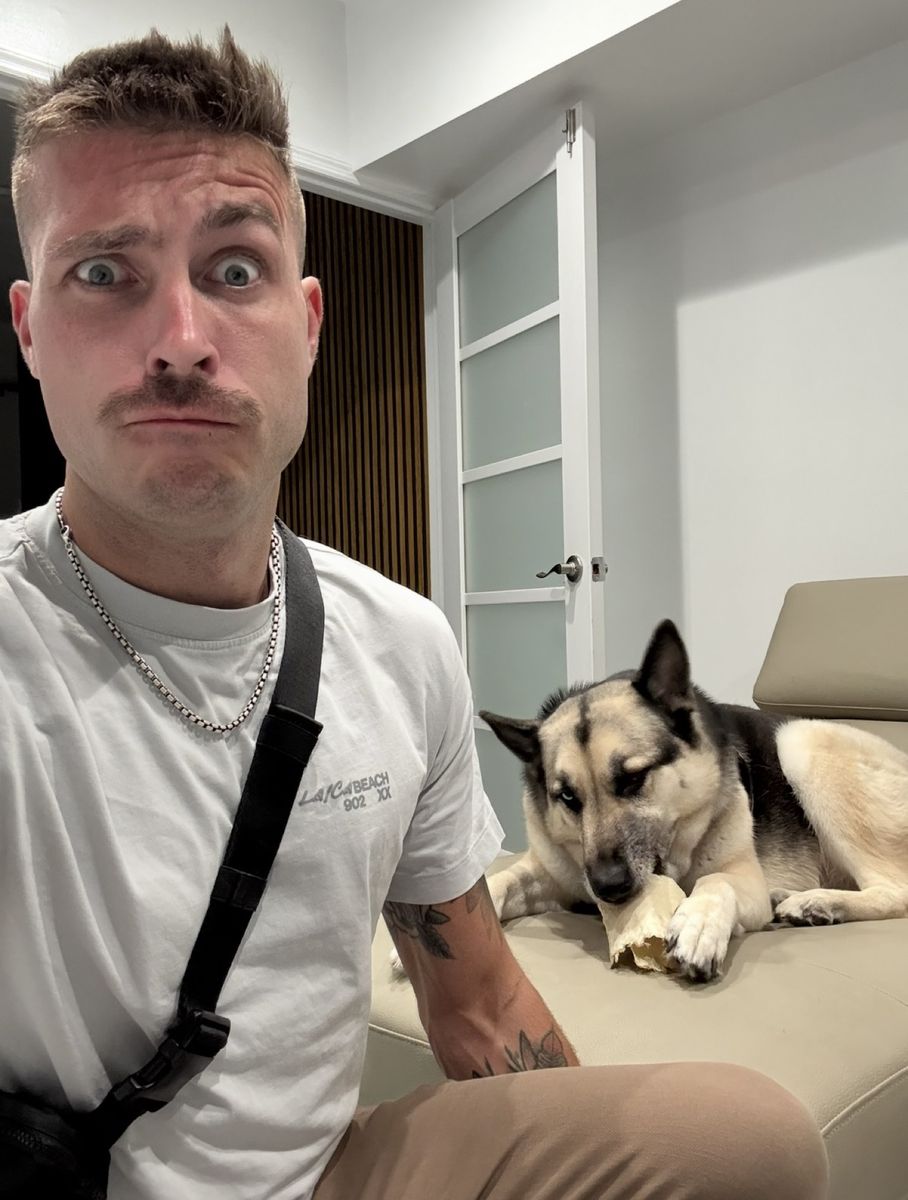Nick pulling a funny wide-eyed face in a white tee while Finn chews a bone on the couch behind him